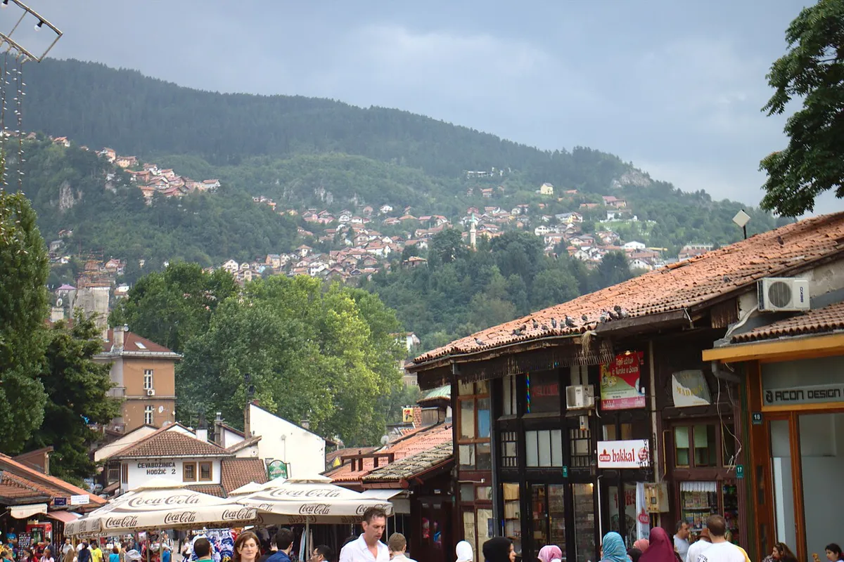 A lively summer street in the Bascarsija bazaar in Sarajevo, Ottoman shopfronts and cafe parasols below a forested hillside of red-roofed houses