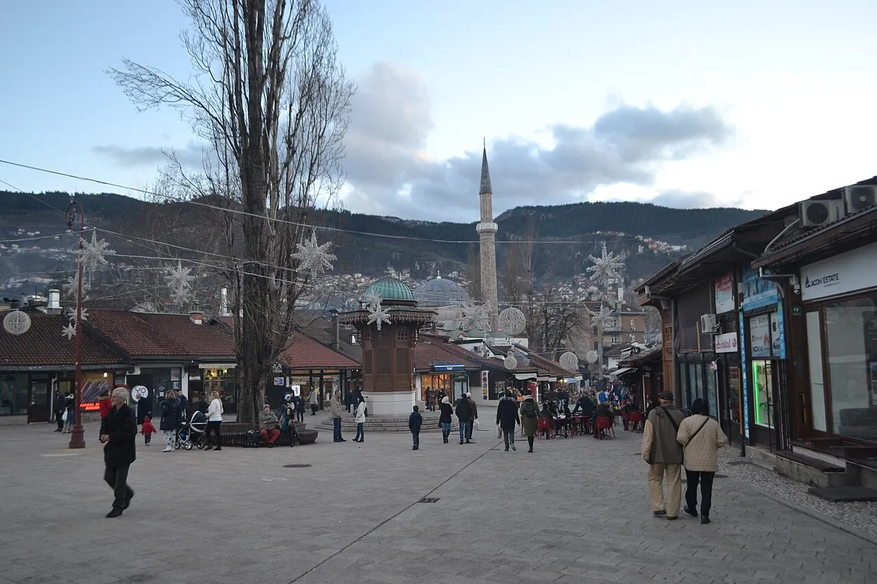 The wooden Sebilj fountain kiosk on Bascarsija square in Sarajevo, a minaret and mosque dome behind, festive lights strung across the square