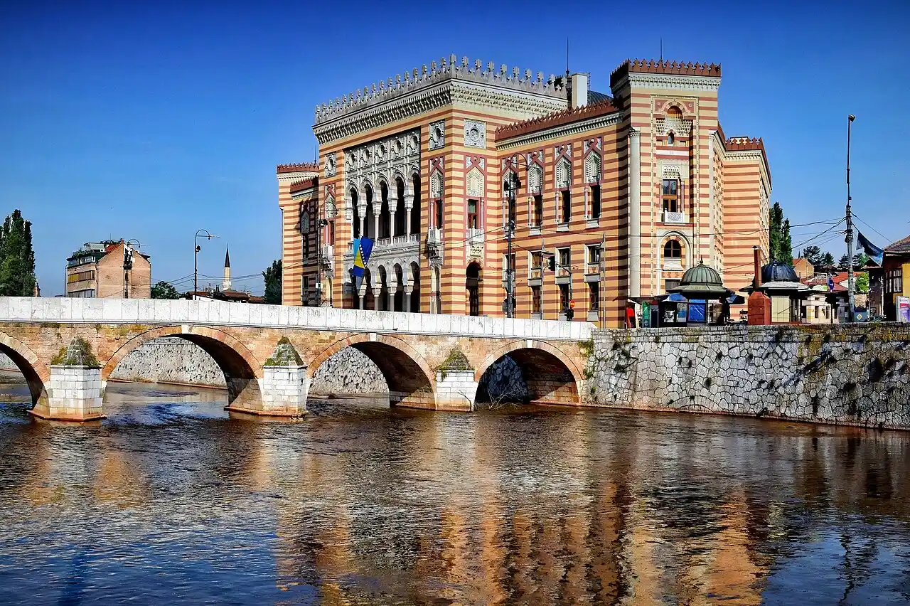 The striped orange and cream facade of Sarajevo City Hall (Vijecnica) beside a stone bridge over the Miljacka river under blue sky