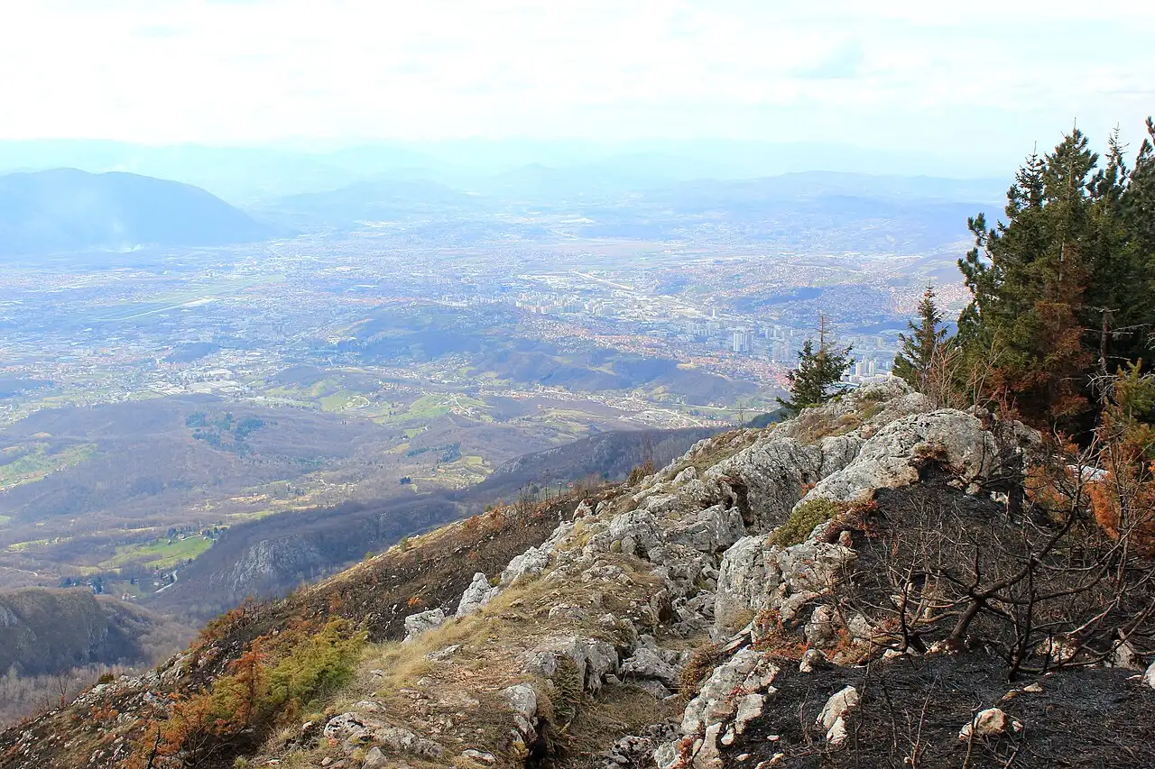 Sarajevo spread across its valley seen from high on Mount Trebević, rocky ground in the foreground and hills beyond
