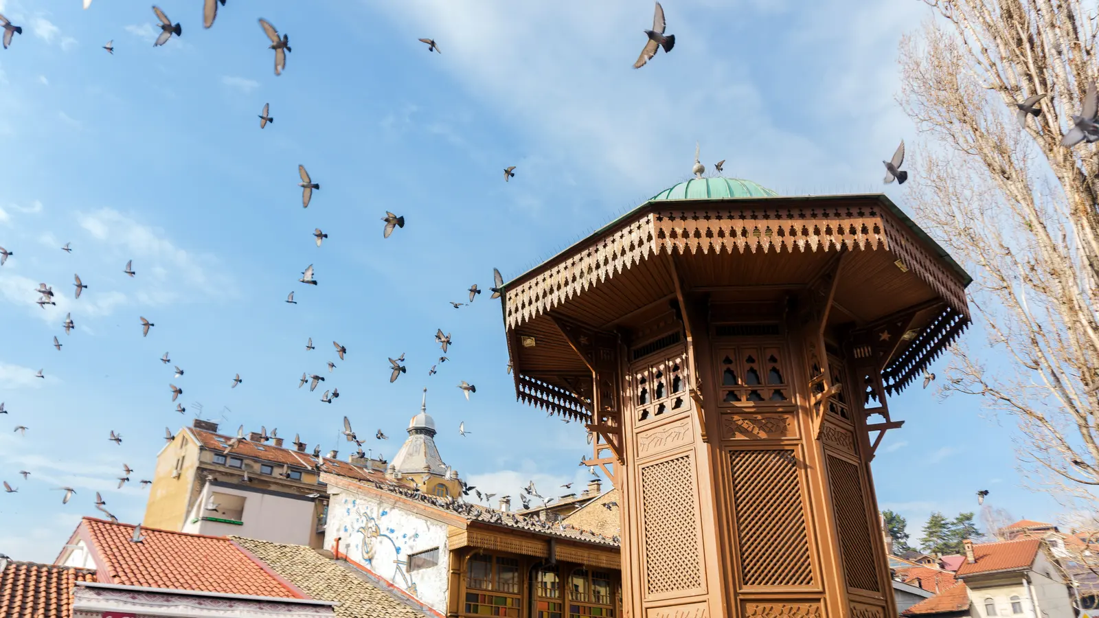The wooden Sebilj fountain kiosk with its green dome, pigeons wheeling overhead against a blue sky in Sarajevo's Baščaršija