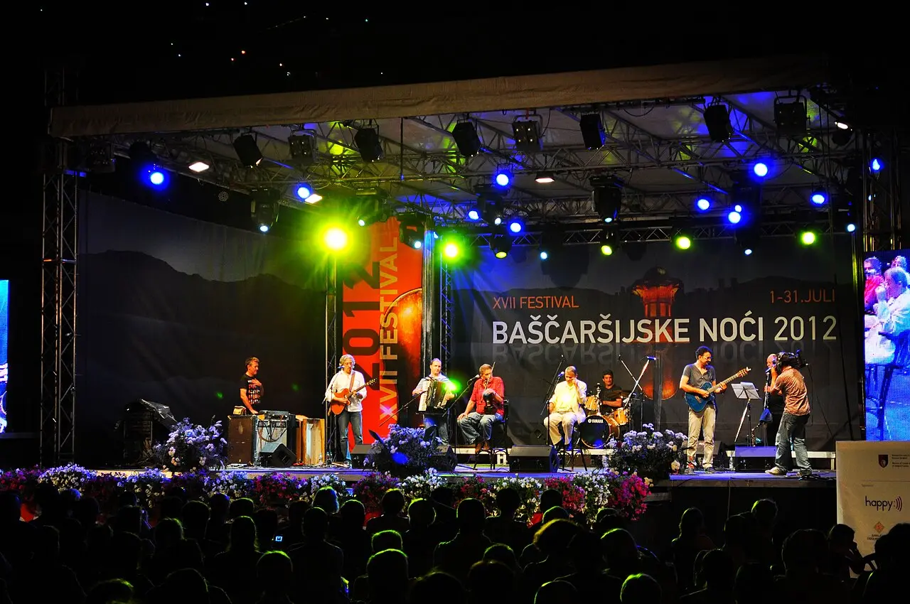 A band playing on a lit open-air stage at night in front of a packed crowd, the backdrop reading Baščaršijske noći with the July dates