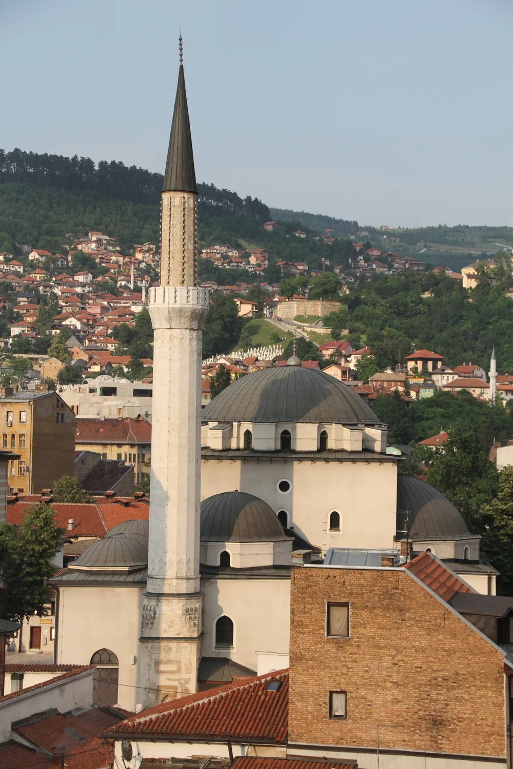 The lead-grey domes and tall stone minaret of the Gazi Husrev-beg Mosque above the rooftops of Sarajevo, the wooded hillside behind