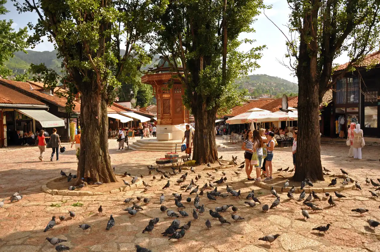 The Sebilj wooden fountain on Baščaršija Square, ringed by pigeons on the cobbles, old bazaar shops and café parasols behind
