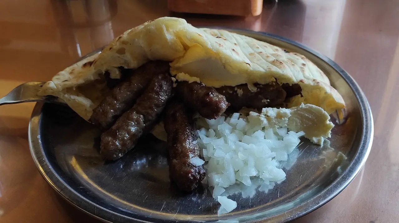 A portion of grilled cevapi tucked into somun flatbread on a metal plate with chopped raw onion and a scoop of kajmak