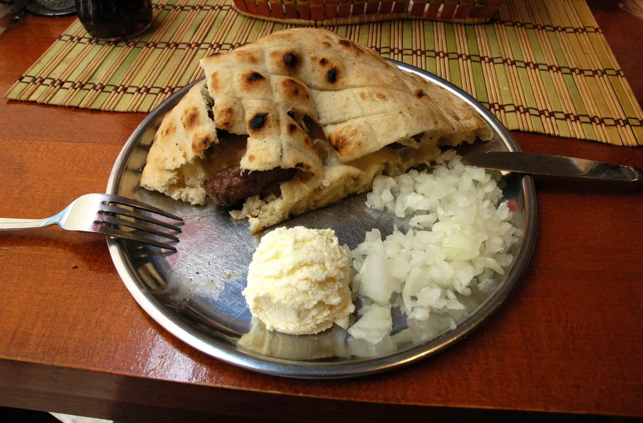 Cevapi in somun on a metal plate with a scoop of kajmak and a pile of chopped onion beside it