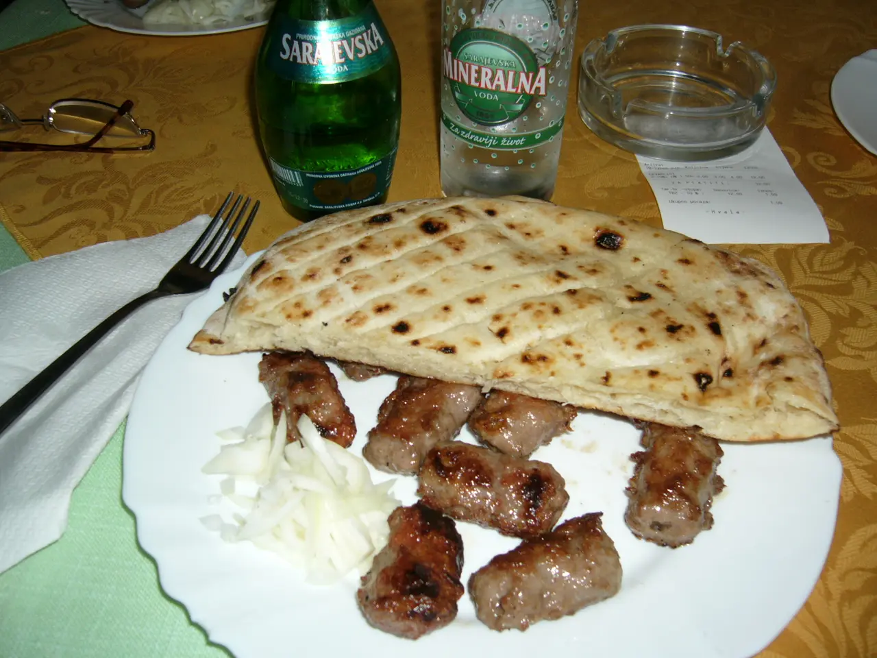 A plate of cevapi with somun and chopped onion on a table with Sarajevska mineral water bottles and the paper bill