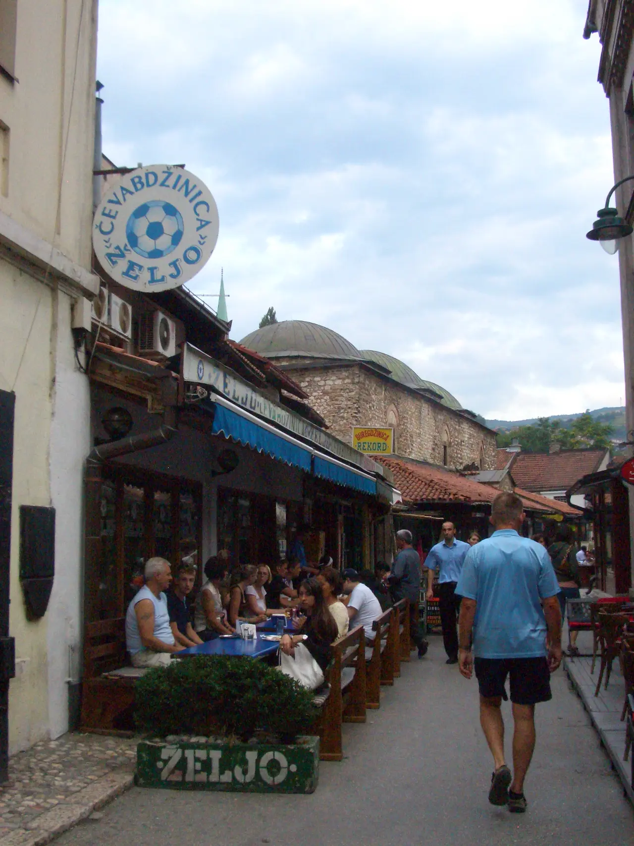 The Cevabdzinica Zeljo shopfront in Sarajevo with a round sign showing a blue and white football, tables along the narrow bazaar lane