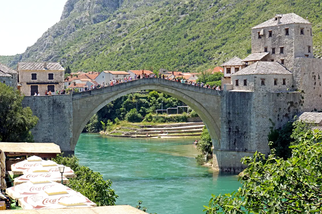 The reconstructed Old Bridge of Mostar arching over the turquoise Neretva, crowds crossing on a hot clear summer day