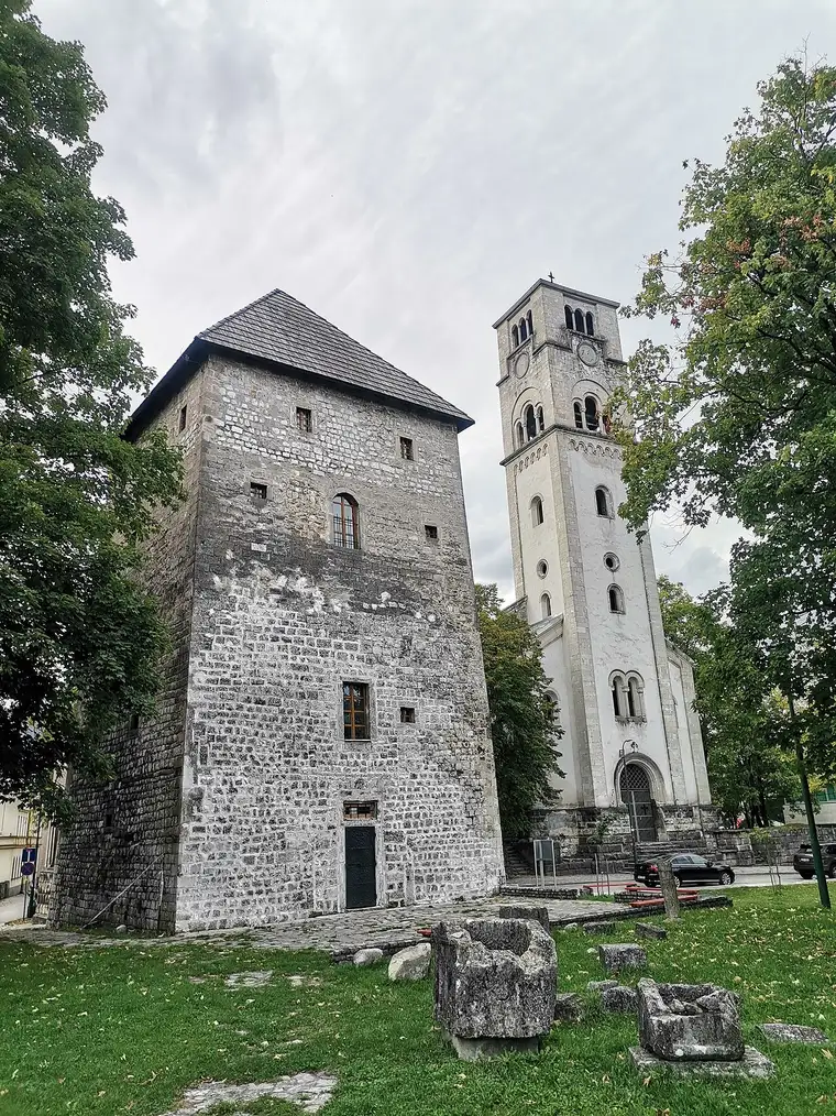 The square stone Captains Tower in Bihac with the tall pale bell tower of St Anthonys church rising behind it and cut stone blocks scattered on the grass