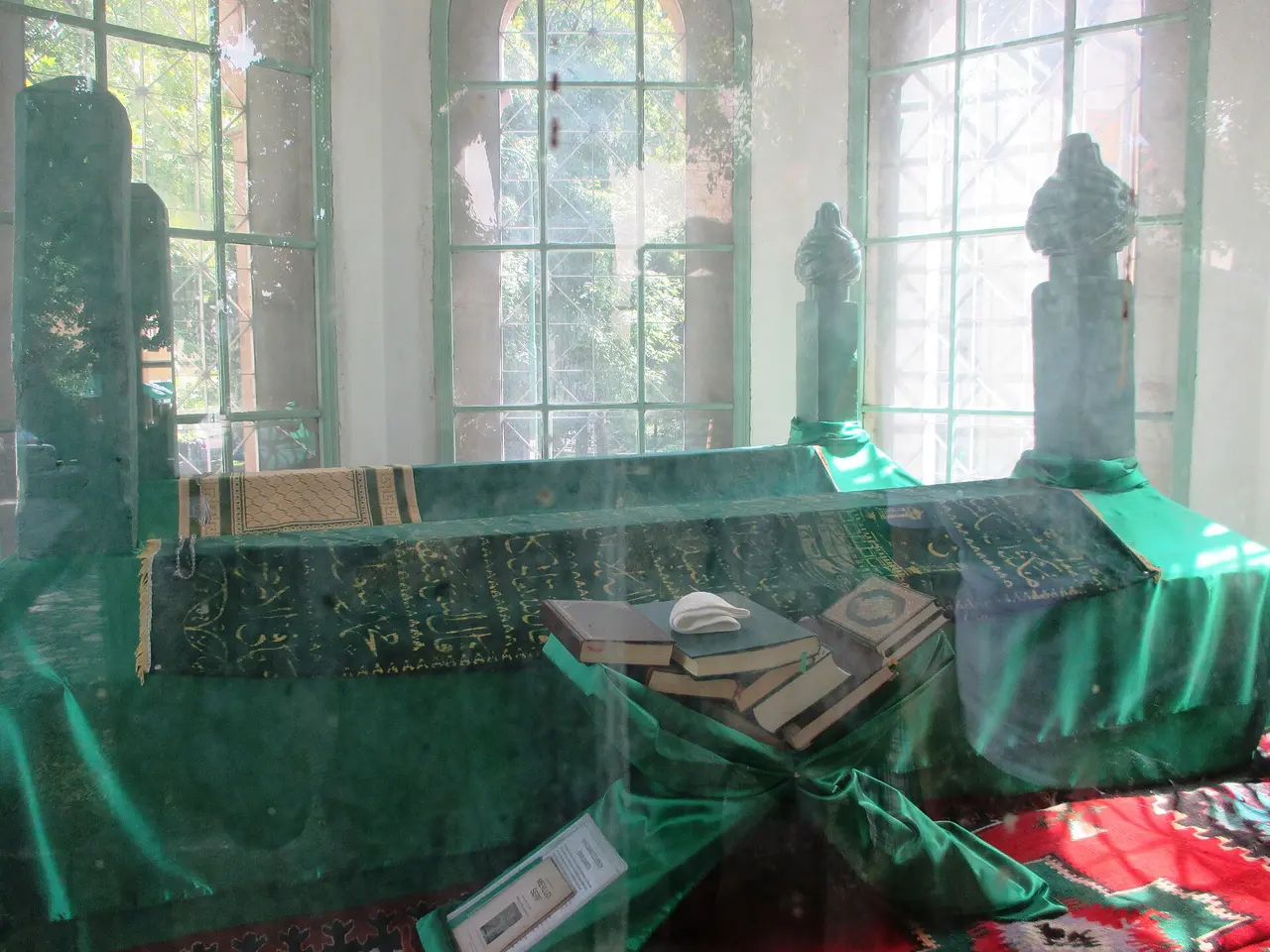 Inside the Turbe mausoleum in Bihac: two long tombs draped in green cloth with gold Arabic script, carpets on the floor and arched windows behind