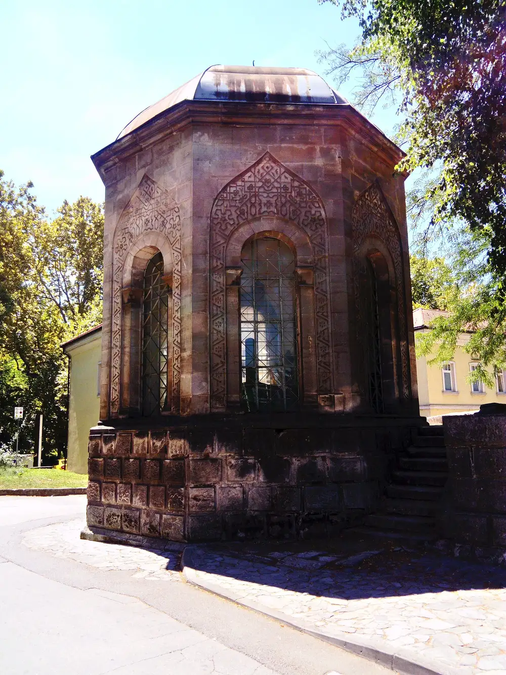 The octagonal stone Turbe mausoleum in Bihac with carved arches and glazed windows, standing beside the road