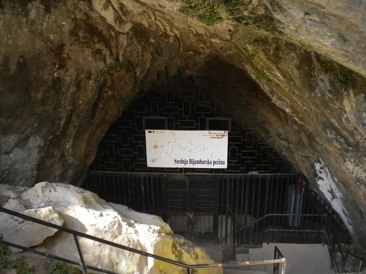 The gated mouth of the Middle Bijambare Cave under a pointed limestone arch, a park sign reading Srednja Bijambarska pecina hung across the bars and steps leading down to it
