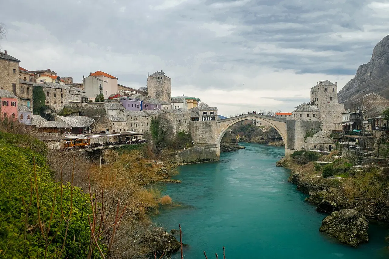 The single-arch Old Bridge of Mostar over the Neretva under a cloudy sky, stone houses and a minaret alongside