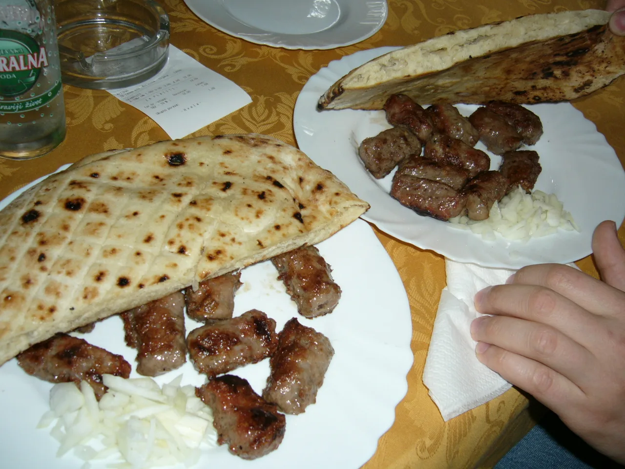 Two plates of grilled ćevapi with somun flatbread and chopped onion, a paper bill on the table next to a bottle of mineral water