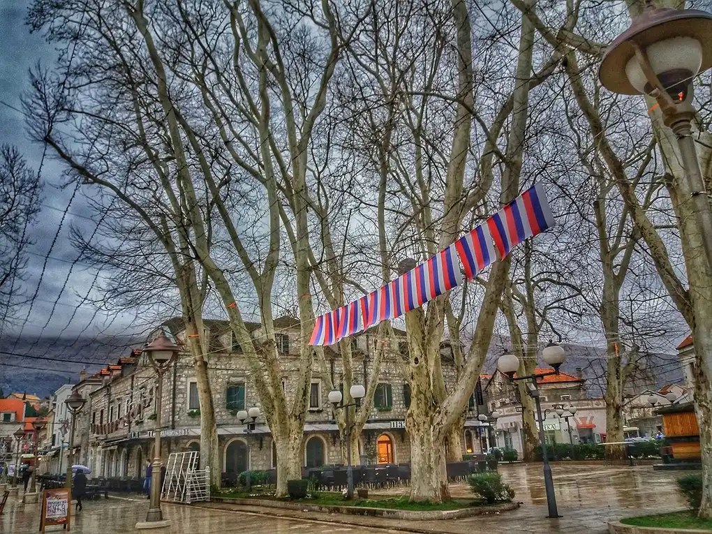 The plane-tree-lined old-town square in Trebinje with café terraces and stone buildings on a wet winter afternoon