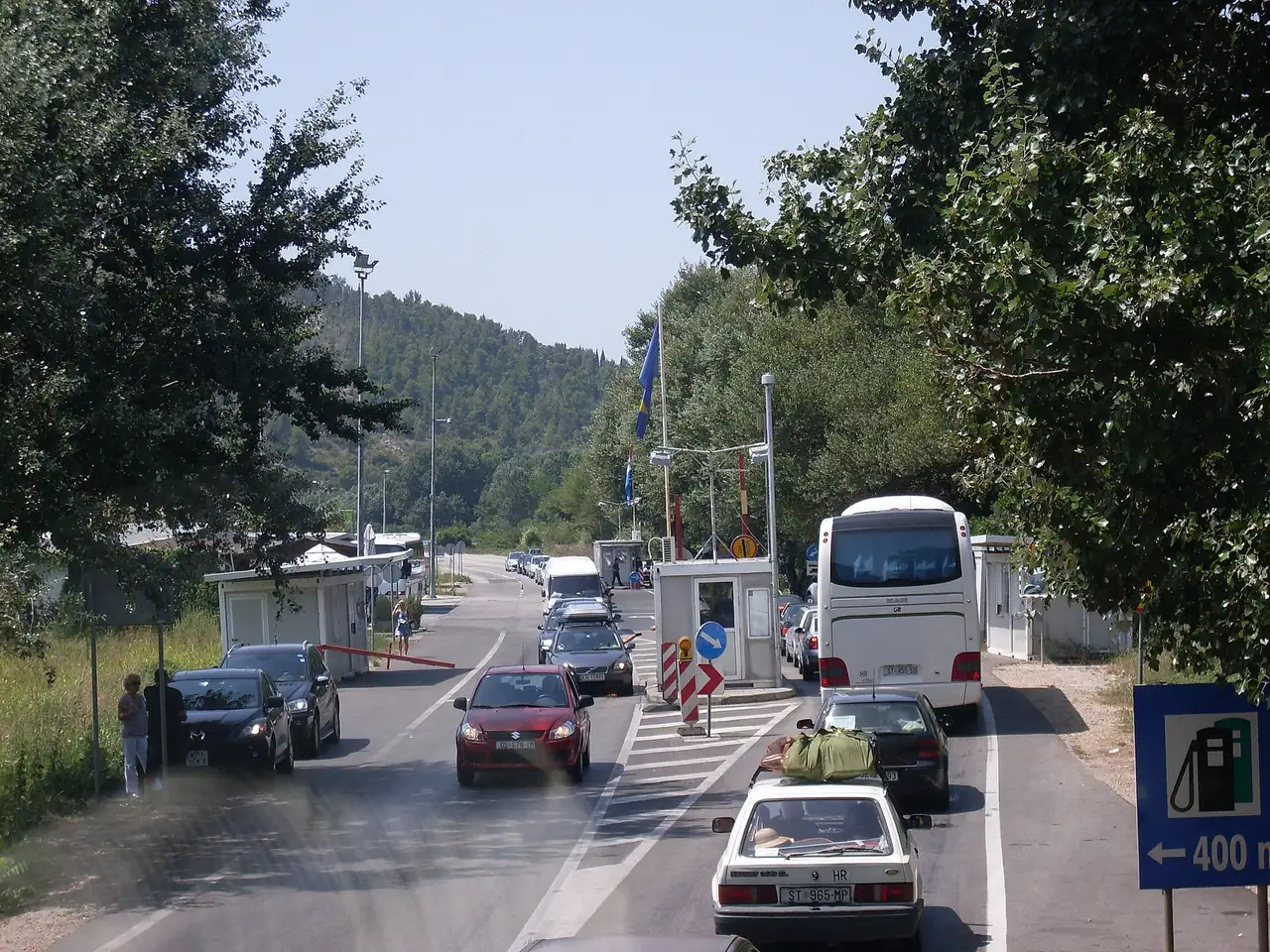 Cars and a coach queuing at the booths of a Croatia-Bosnia road border crossing in summer, a flag flying over the checkpoint
