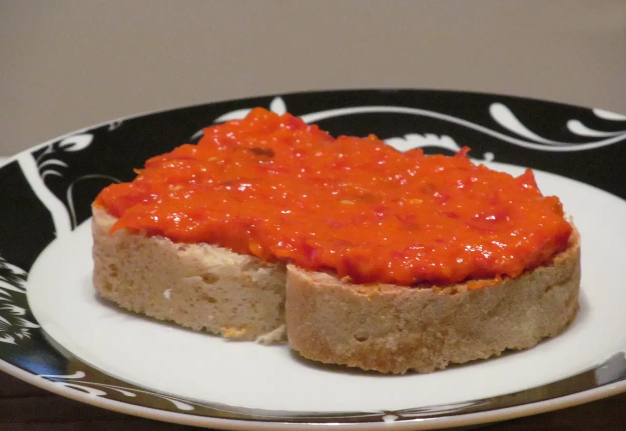 A thick layer of deep red home-made ajvar spread on a slice of bread on a black and white plate