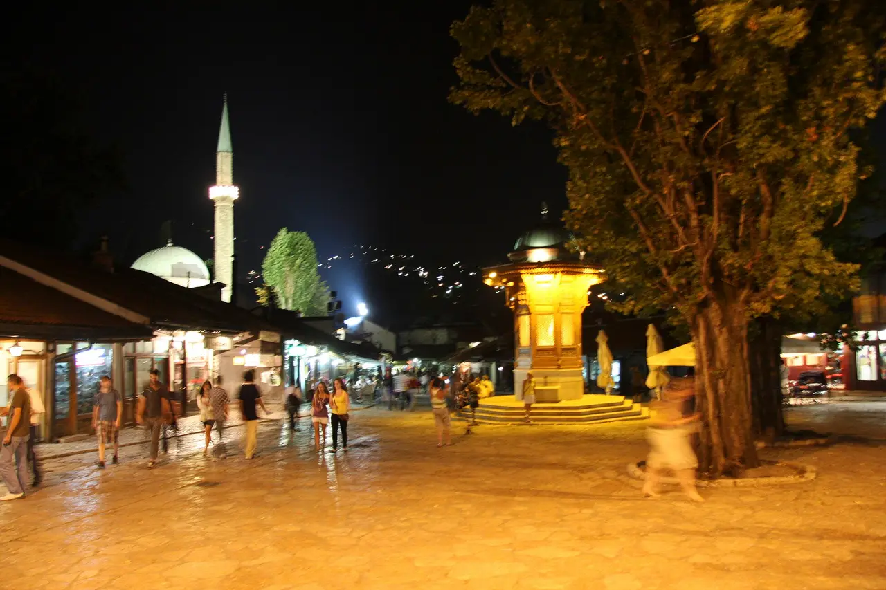 The lit Sebilj fountain and shopfronts of Bascarsija in Sarajevo on a warm summer night with people crossing the square