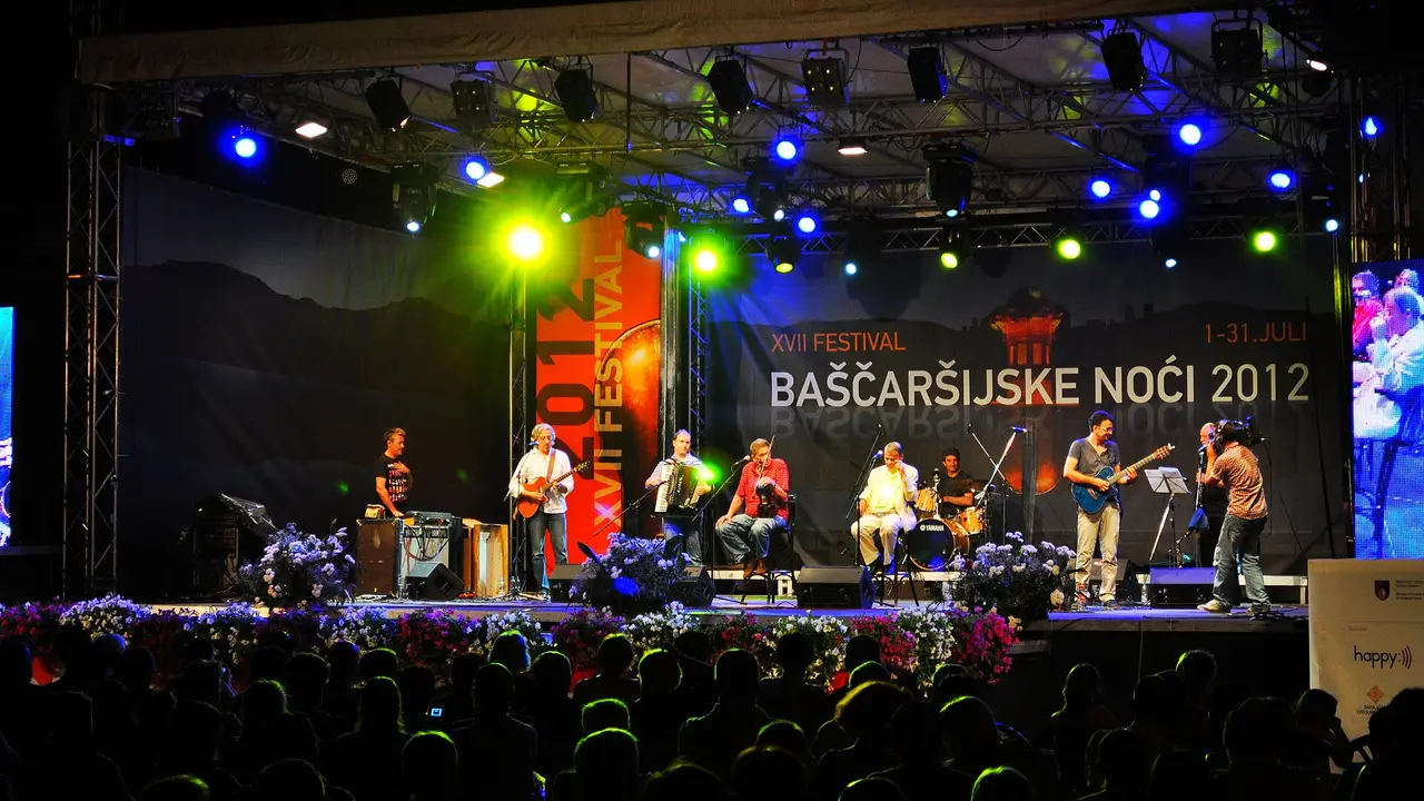 A lit outdoor stage at the Bascarsija Nights festival in Sarajevo, a band of musicians playing in front of a large festival banner and a dark crowd of heads in the foreground