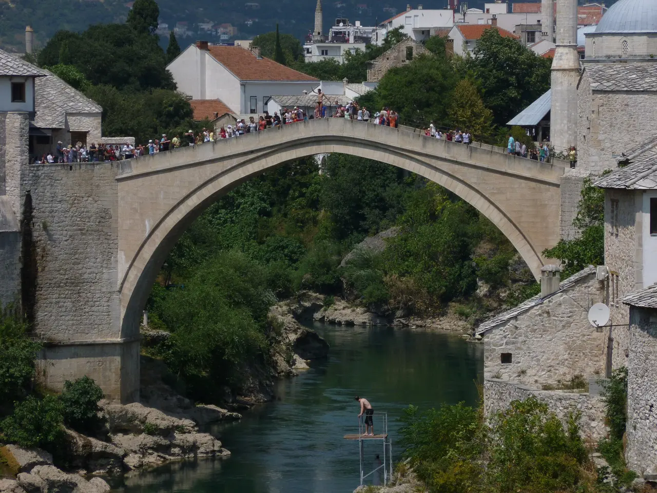 Crowds lining the arch of the Old Bridge in Mostar in summer while a diver stands on a small platform above the green Neretva below