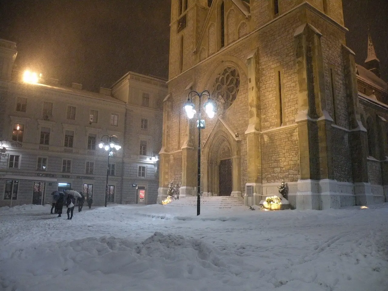 Sacred Heart Cathedral in Sarajevo lit up at night during heavy snowfall, deep snow across the square and a few people walking under umbrellas