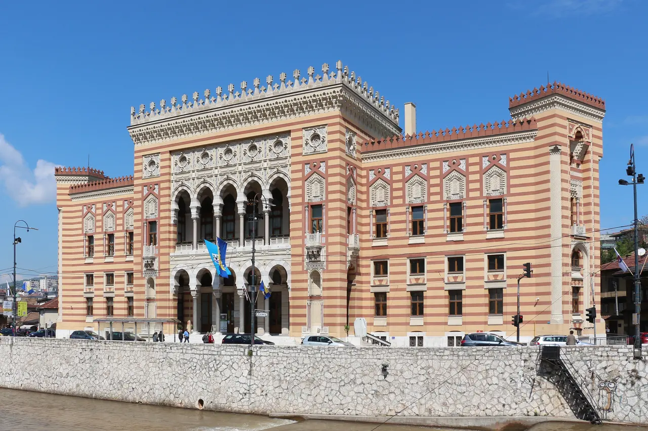 The striped Moorish Revival facade of Sarajevo City Hall (Vijecnica) beside the Miljacka river under a clear sky