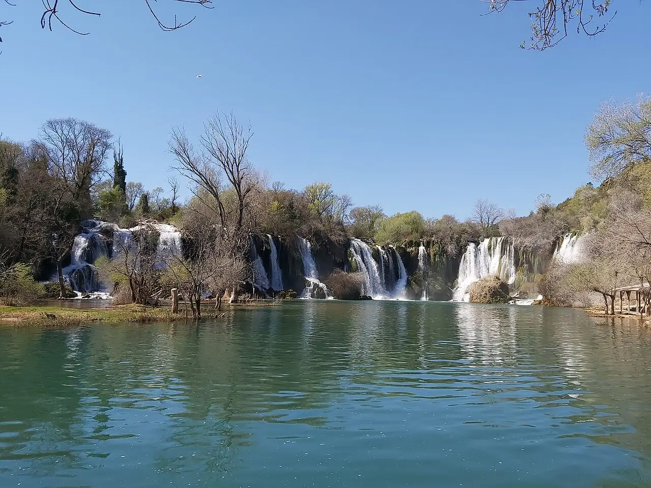 The Kravice waterfalls running full over a wide travertine ledge into a turquoise pool, bare leafless trees on the banks in the cool season