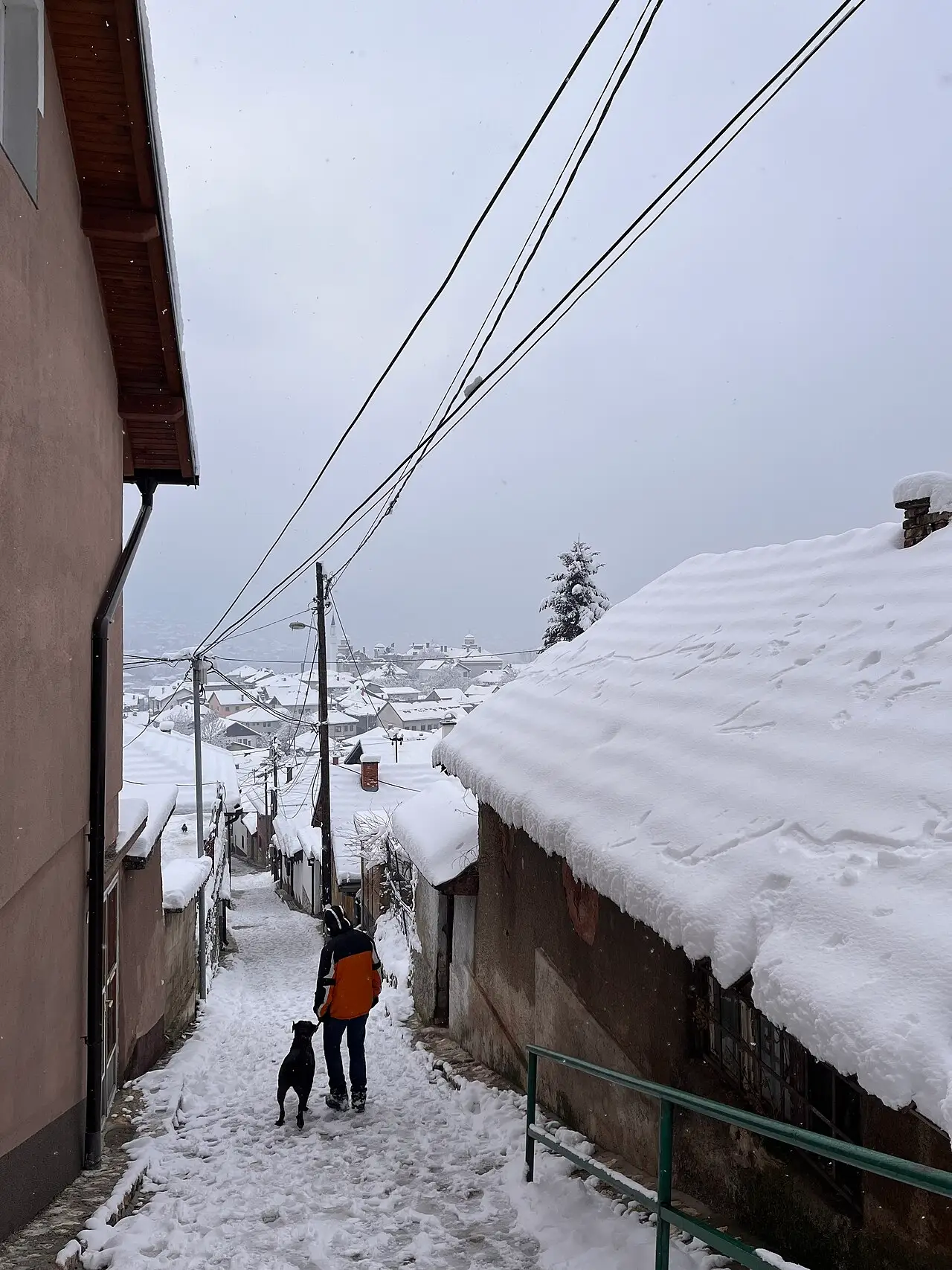 A person walking a dog down a snowy cobbled lane in the old Vratnik quarter of Sarajevo, snow heaped on Ottoman-era rooftops