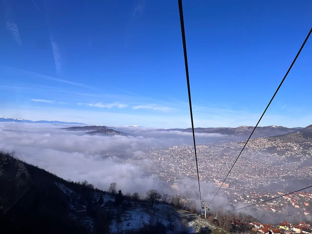 A cable-car view over Sarajevo lying under winter mist in its valley, snow-dusted hills all around under a blue sky
