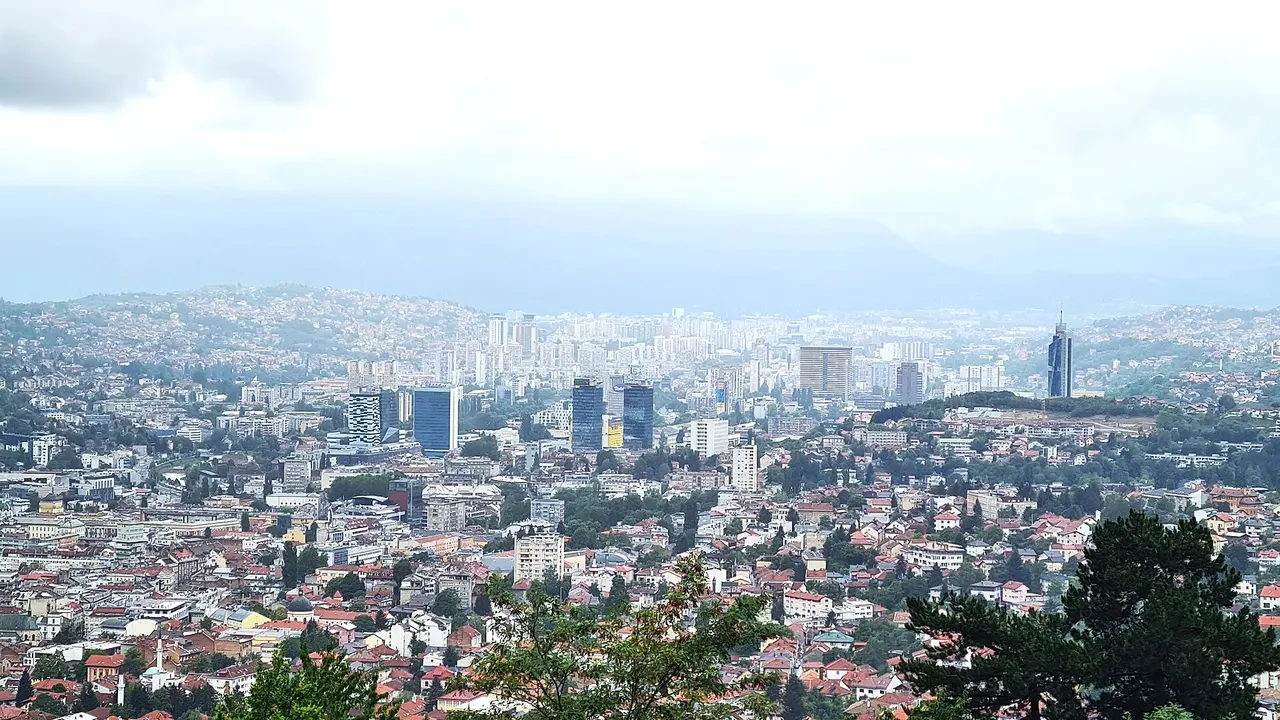 Sarajevo filling its long valley at dusk, minarets and apartment blocks under the surrounding green hills