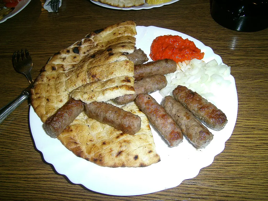 A plate of grilled ćevapi with flatbread, chopped onion and red ajvar on a Bosnian restaurant table
