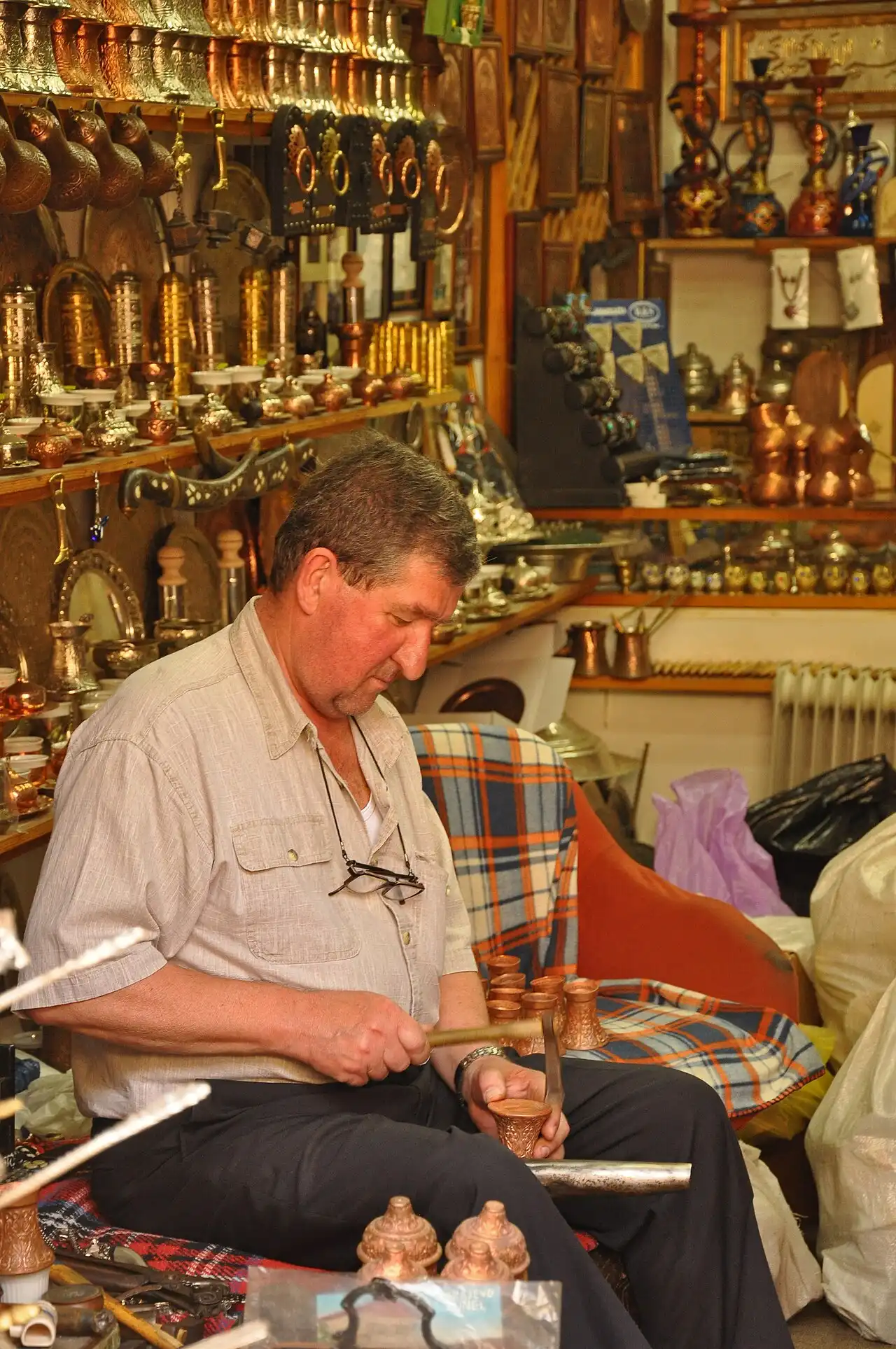 A coppersmith hammering a small coffee cup in his workshop hung with copper trays and pots in Sarajevo