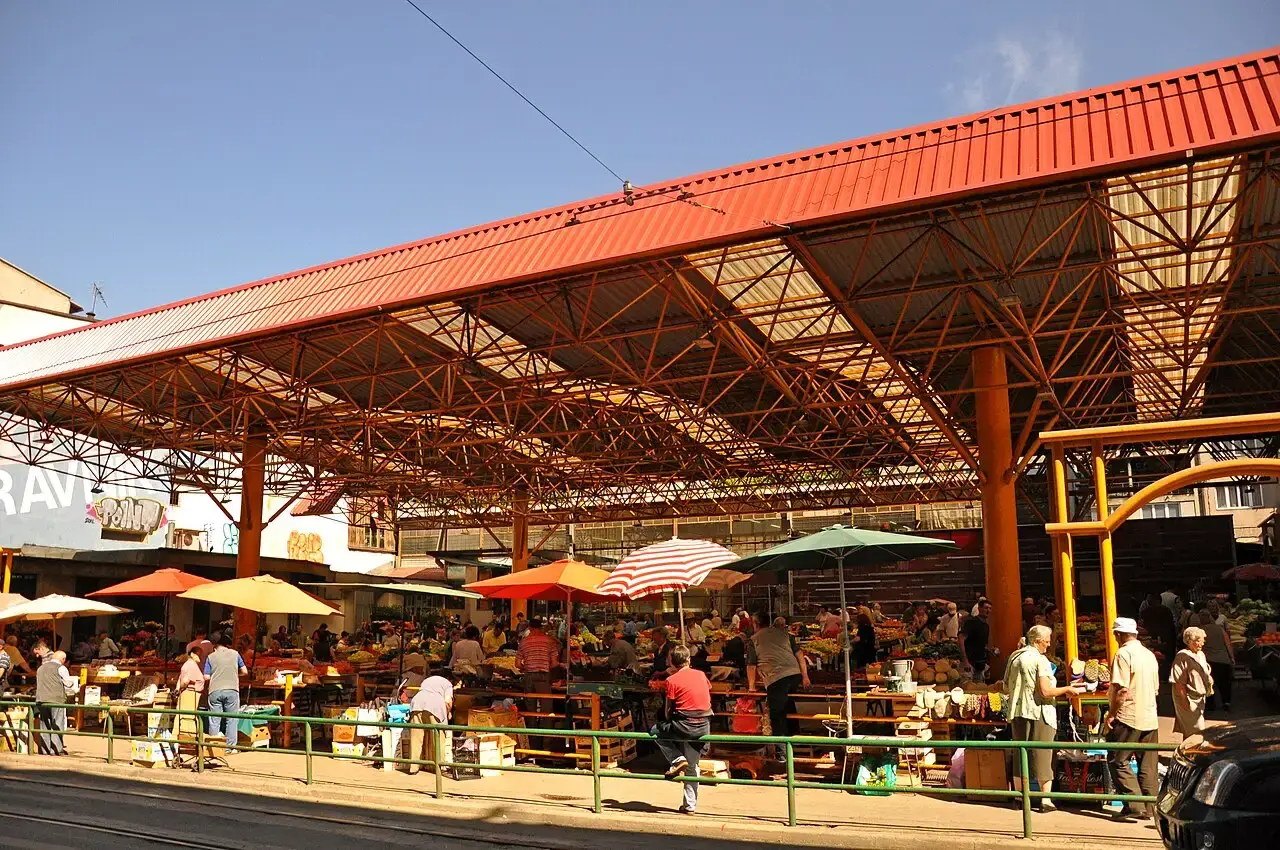 Shoppers under the red roof of an open-air produce market in Sarajevo, stalls piled with fruit and vegetables