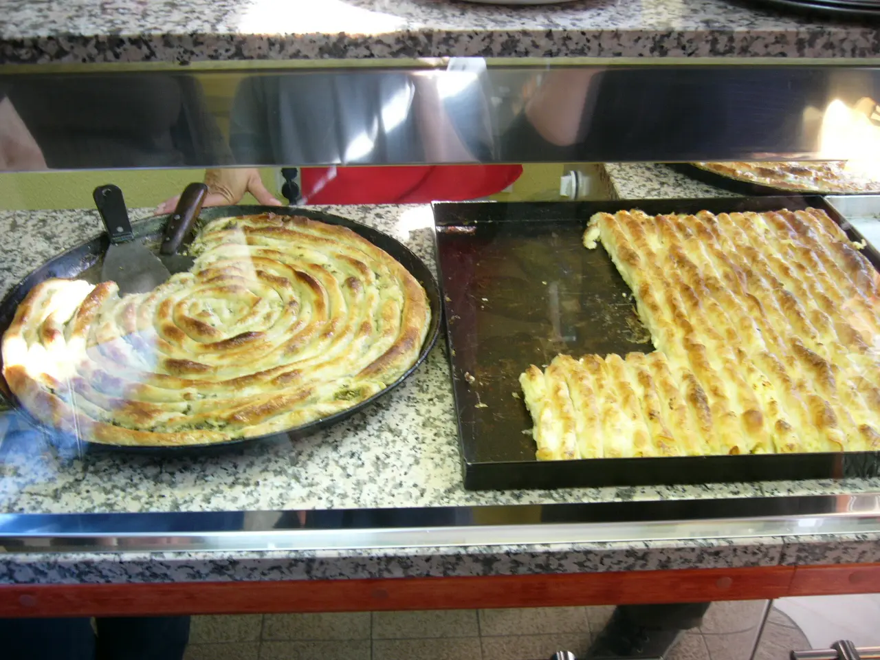 A buregdzinica display counter with a round tray of coiled spinach pita beside a square tray of layered cheese pita, a server cutting a slice