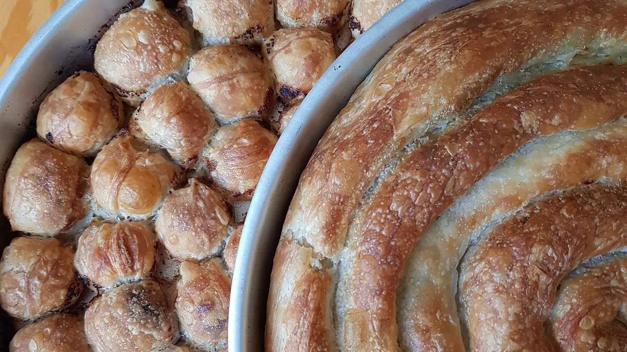 Two round baking trays of Bosnian pita fresh from the oven, one packed with small rolled coils and one holding a single large golden spiral