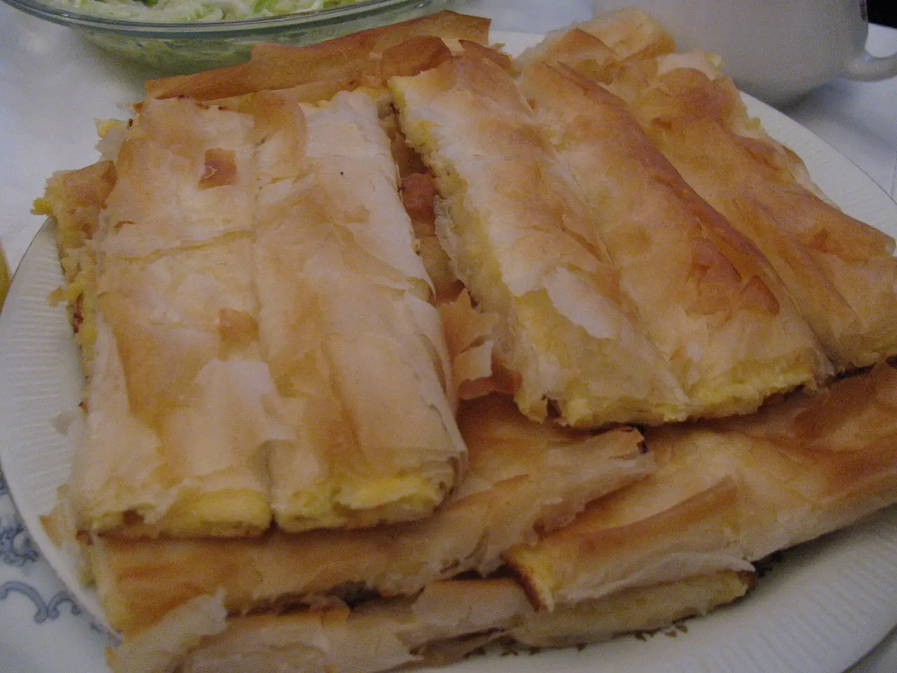 Pale flaky strips of sirnica cheese pita piled on a white plate with a bowl of salad behind