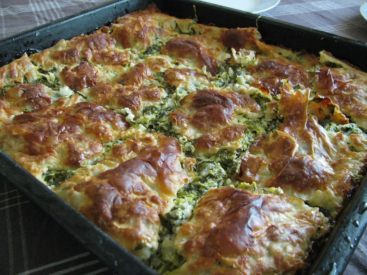 A black baking tray of zeljanica, spinach and cheese pita, browned on top and cut into squares