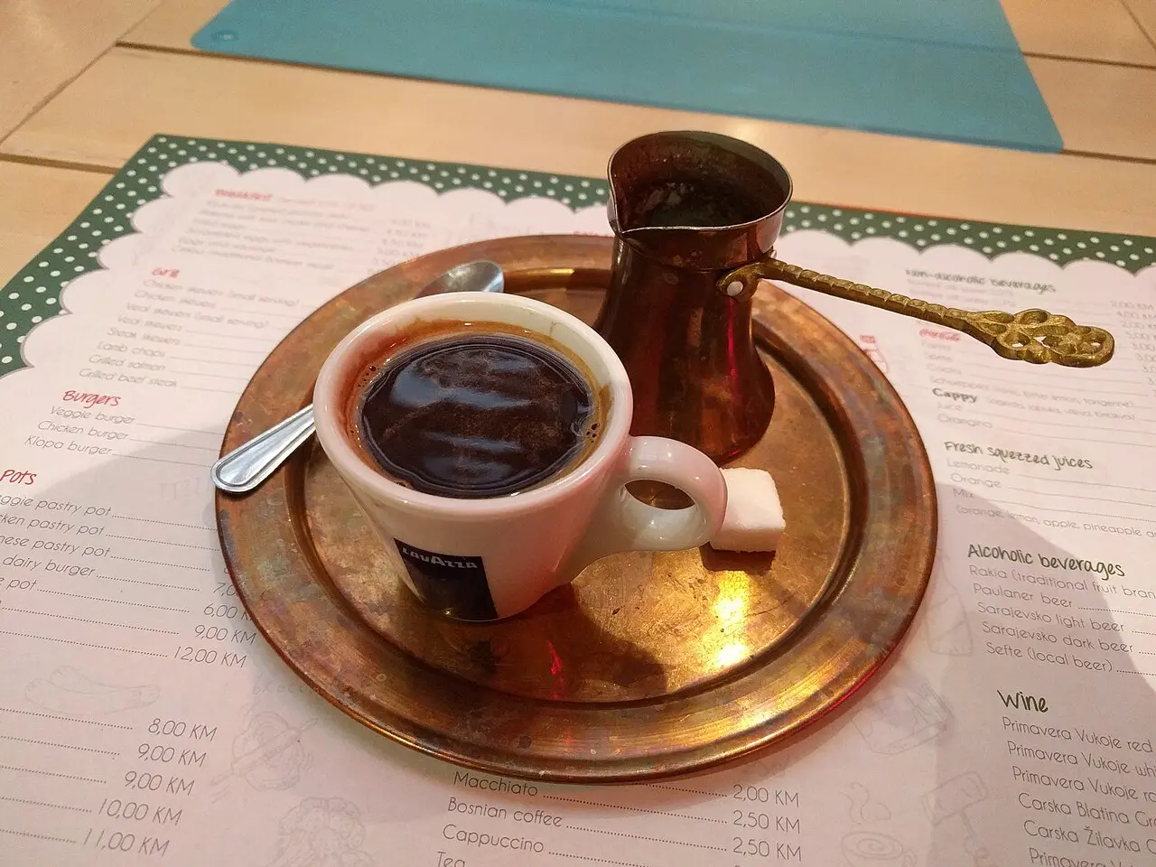 A cafe table in Bosnia with coffee in a copper džezva and cup on a copper tray, resting on a menu that lists Bosnian coffee at 2.50 KM