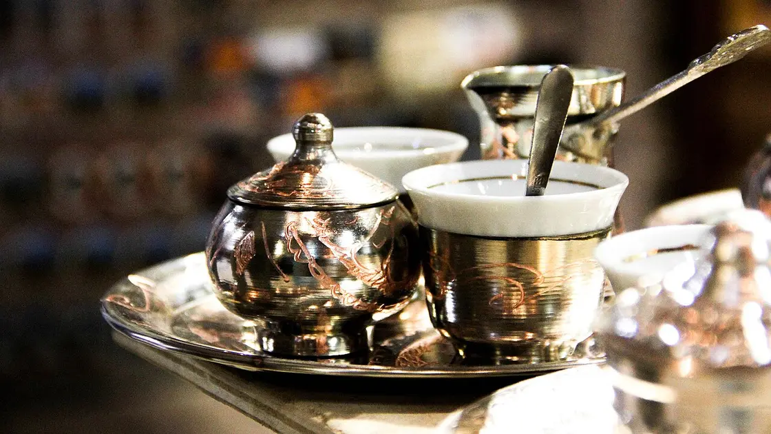 A Bosnian coffee set in a warm cafe: a copper džezva pot, a handleless fildžan cup of coffee and a lidded copper sugar bowl on a metal tray