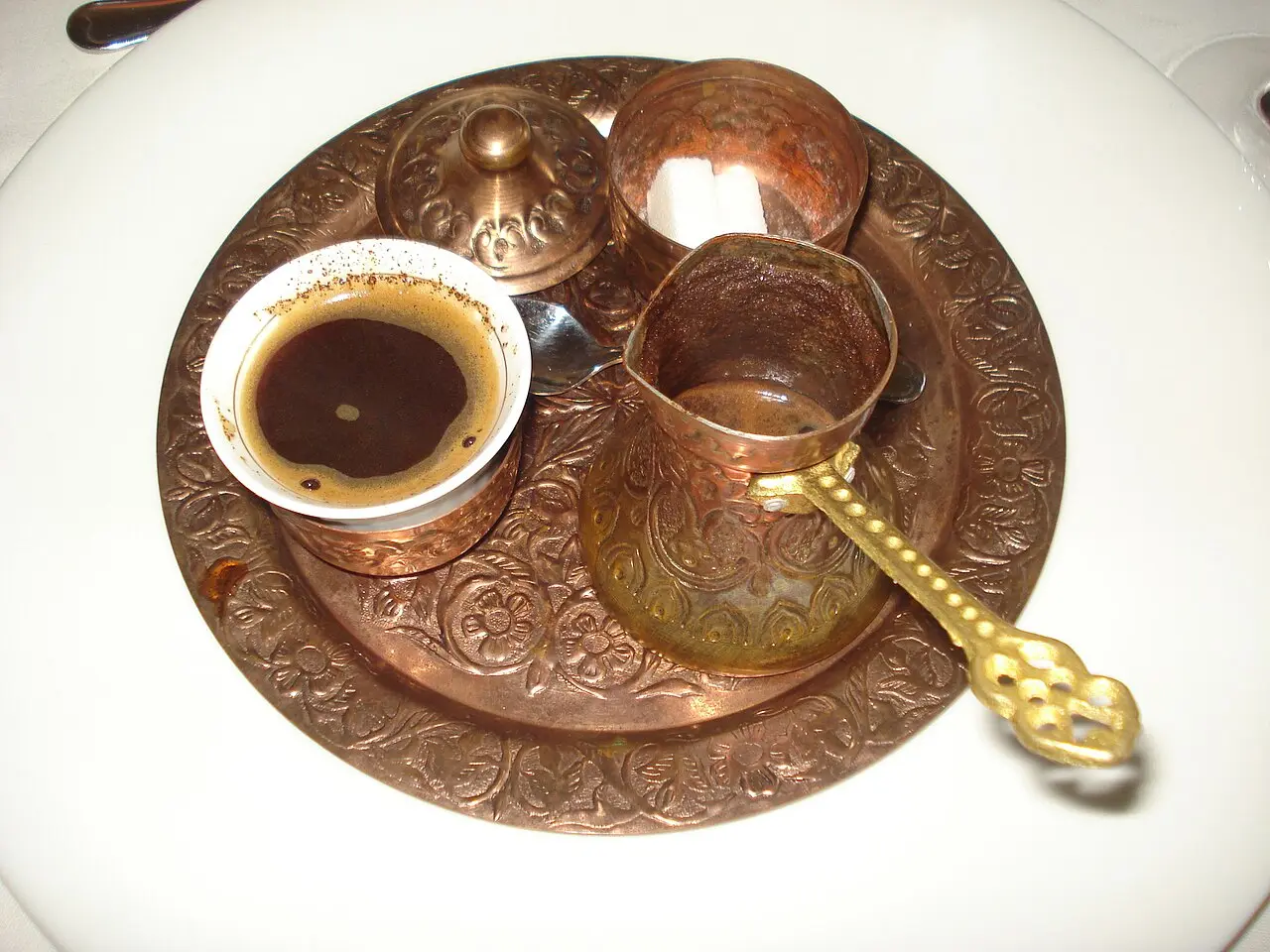 Close view of a fildžan of Bosnian coffee with foam on top, a copper džezva with grounds, a lidded sugar pot and a single sugar cube on an engraved tray