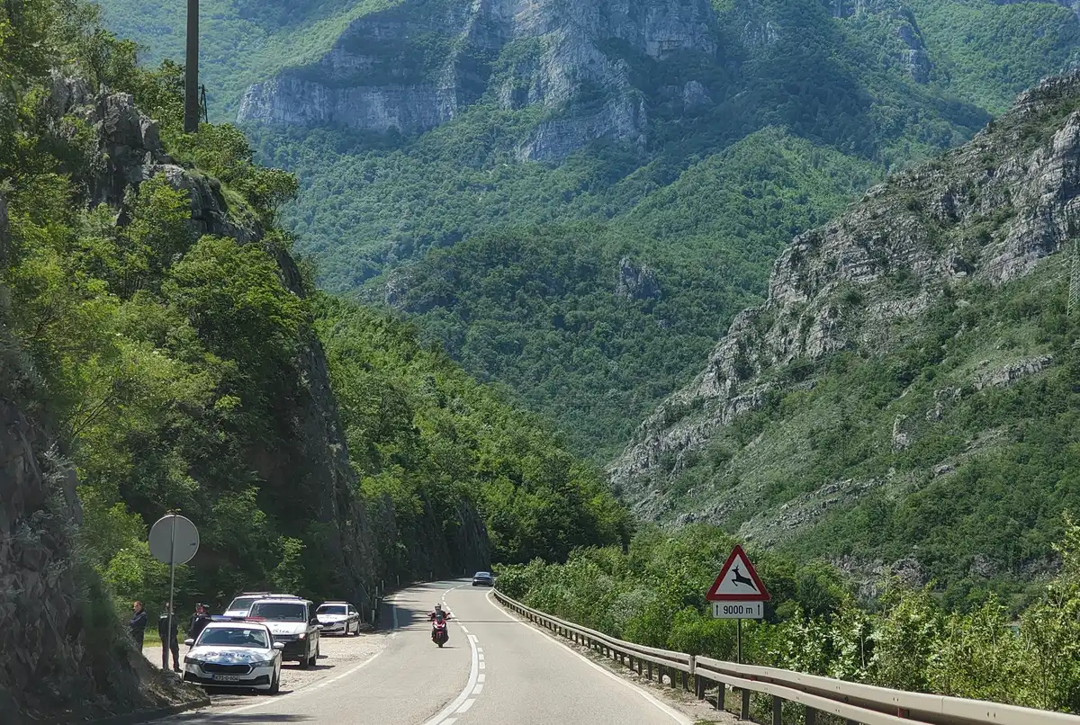 A two-lane road through a steep green gorge in Bosnia with a motorcyclist and police cars parked on the verge