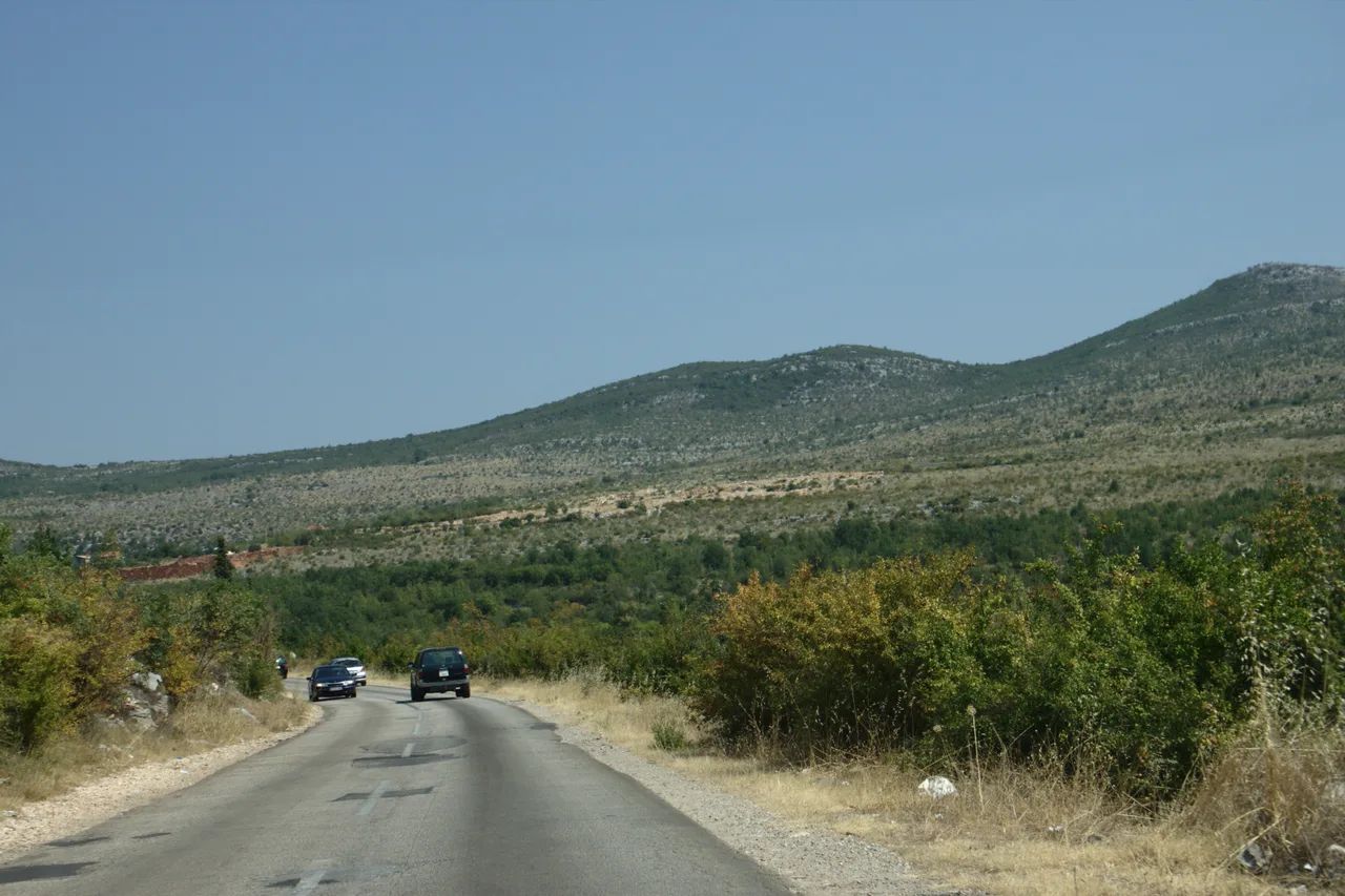 An open two-lane road running through dry karst hills near Kravice in southern Herzegovina, a couple of cars in the distance