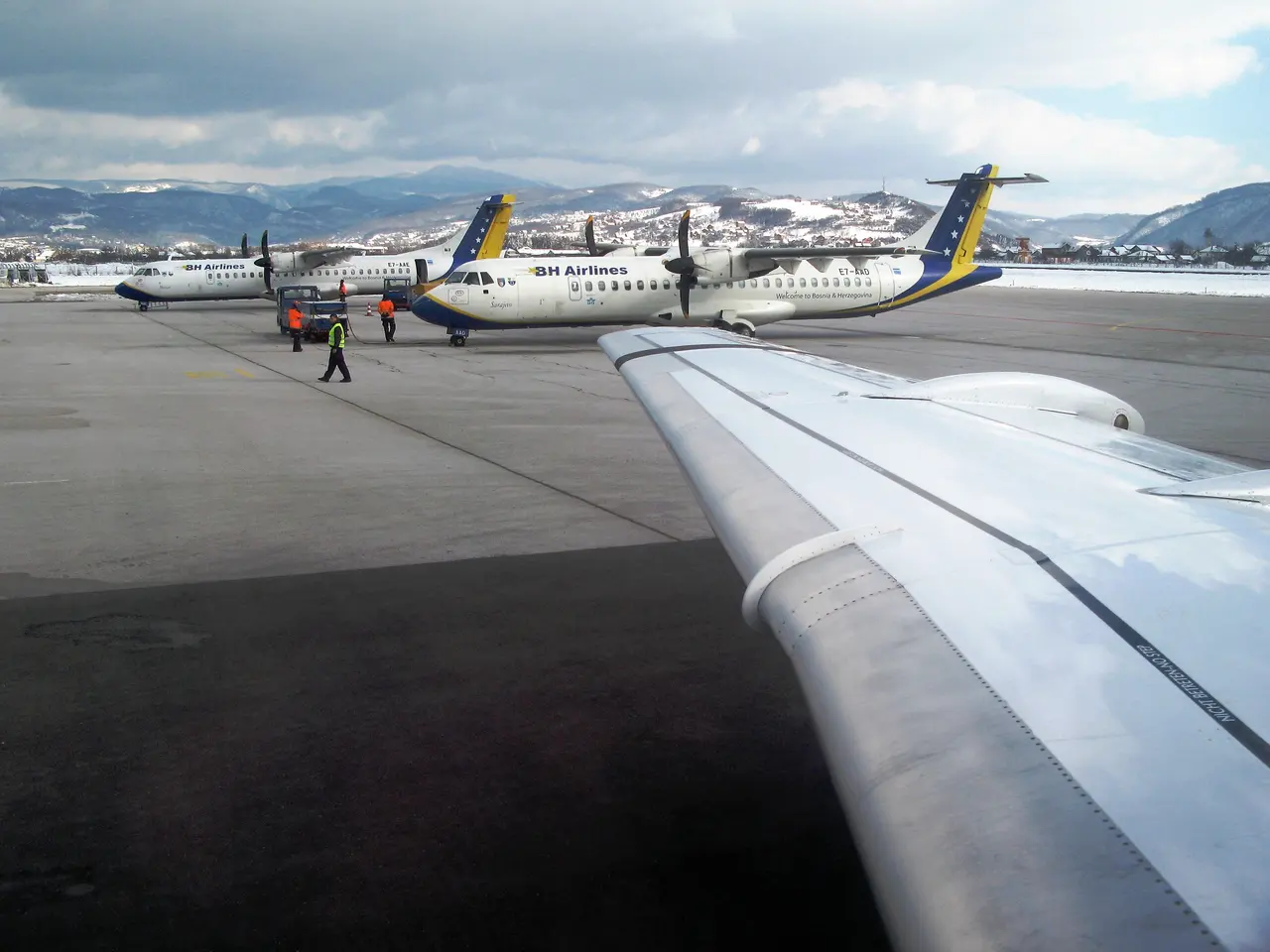 Taxis and parked cars on the forecourt outside the Sarajevo Airport arrivals hall, with hills and a signal tower behind