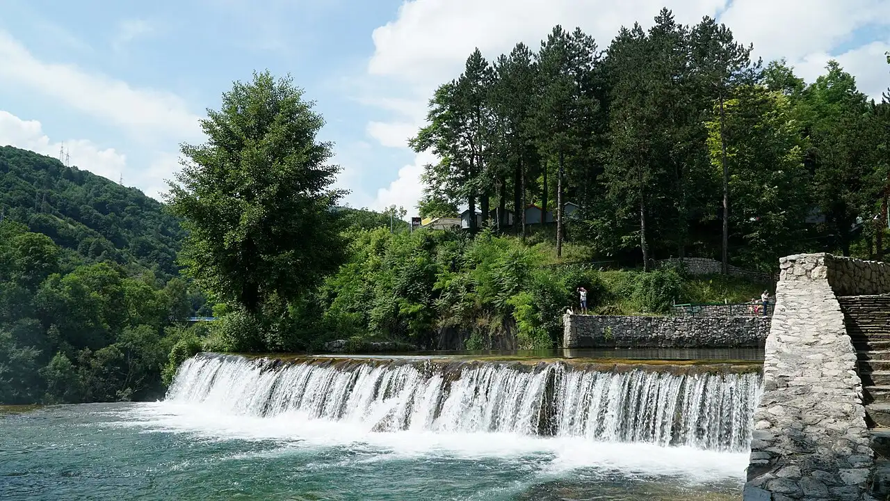 The Pliva Waterfall spreading in a wide white sheet over a green tufa ledge in the centre of Jajce, stone houses on the hill behind