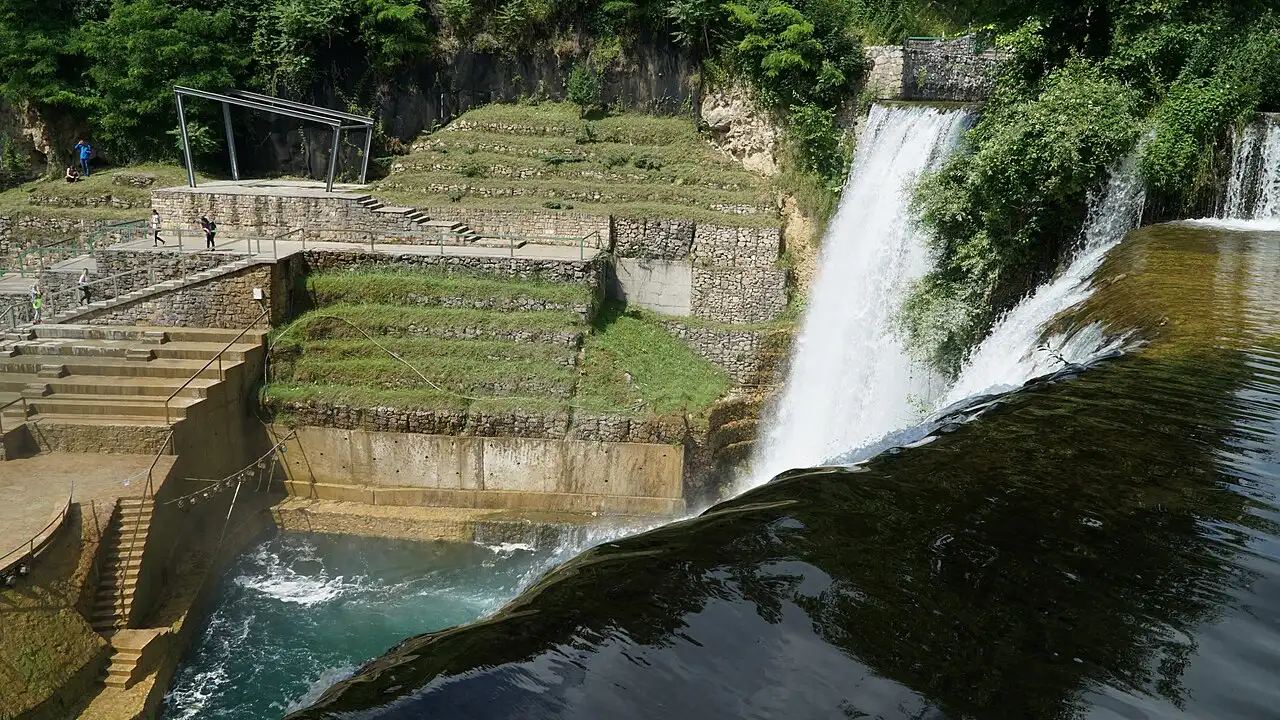 The Pliva Waterfall in Jajce seen from the lower platform, a broad curtain of water with the old town climbing the hill