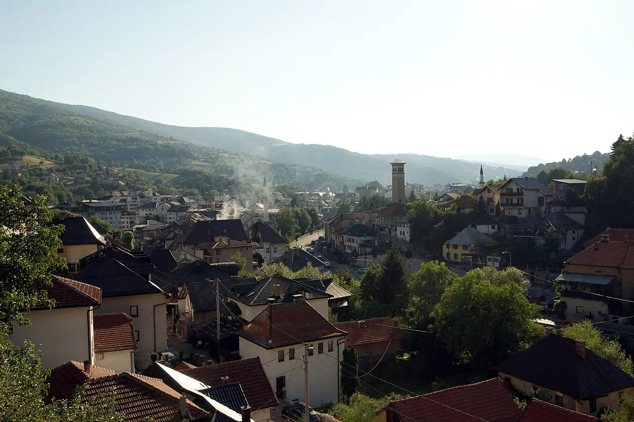 Red roofs and minarets of Travnik filling a green valley under wooded hills, seen from above the old town