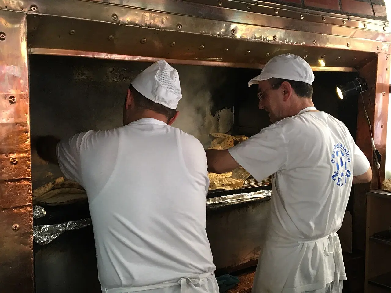 Two cooks in a Sarajevo grill house tending flatbread at a copper-hooded ćevapi oven