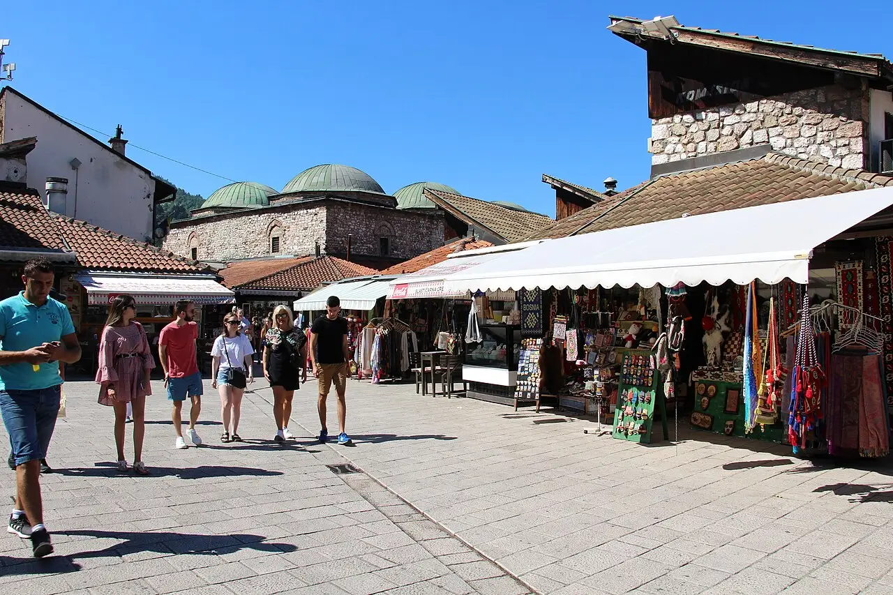People strolling the pedestrian bazaar of Baščaršija in Sarajevo past souvenir stalls with the domed Brusa Bezistan behind