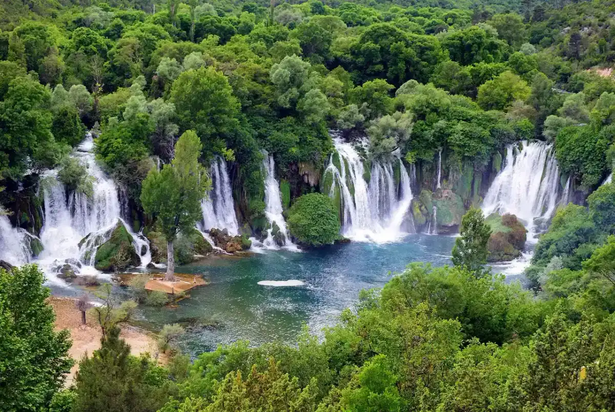 The wide horseshoe of Kravice Waterfalls pouring over a green travertine ledge into a turquoise pool ringed by forest